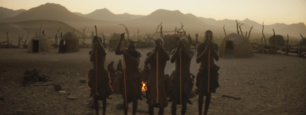Men with sticks dancing with a campfire huts a mountain range the sky and clouds in the background