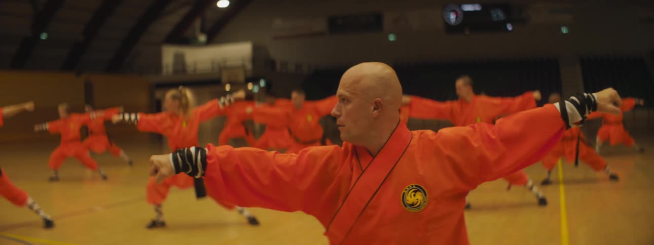 A group of men and women perform martial art in a gym wearing orange outfits