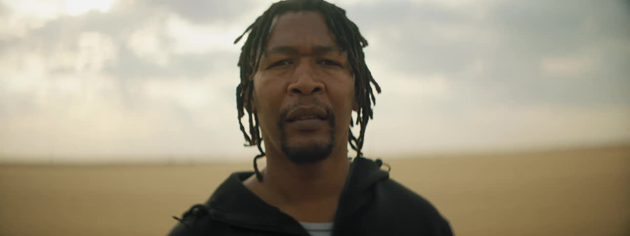 A middle aged black man stares into the camera while standing in a barren desert on a cloudy day