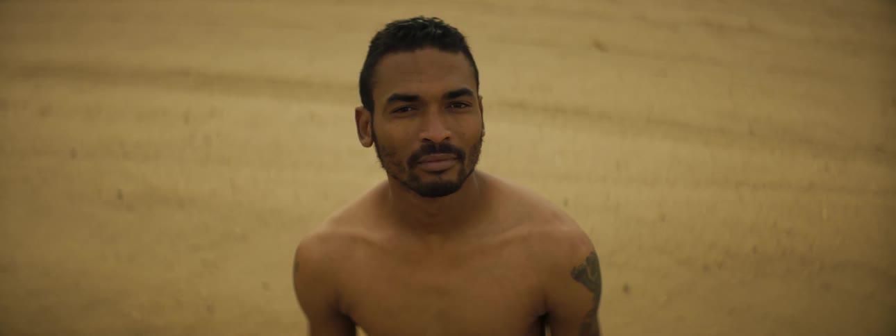 Man with a shoulder tattoo and a beard stands on the sand and looks at the camera on the beach