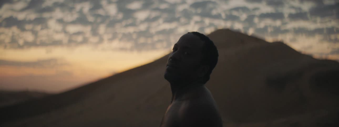 A man in a desert looking up at the sky with a sand dune the sky and clouds in the background