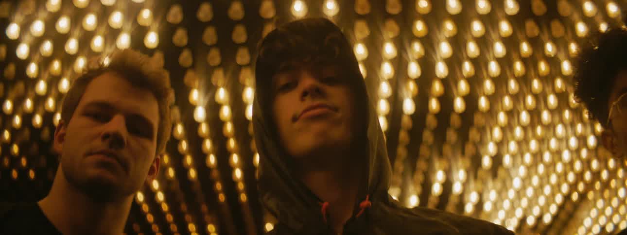 A group of young men nod their heads as they gaze into the camera and pose together under a flashing array of ceiling lights