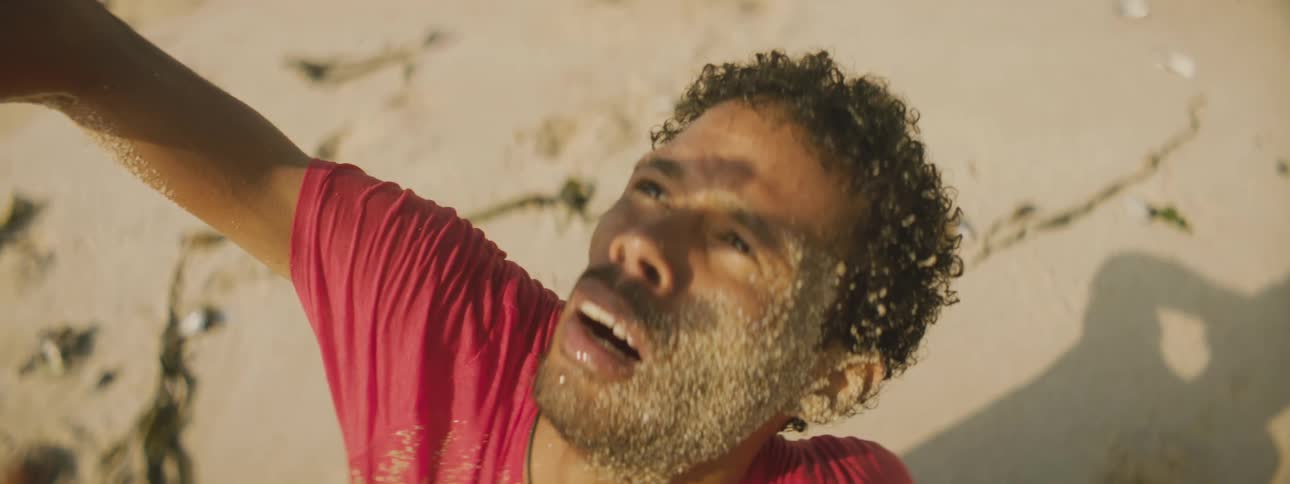 A man covered in sand in a desert holds his hand up and looks up at the sky as another person lies in the sand next to him