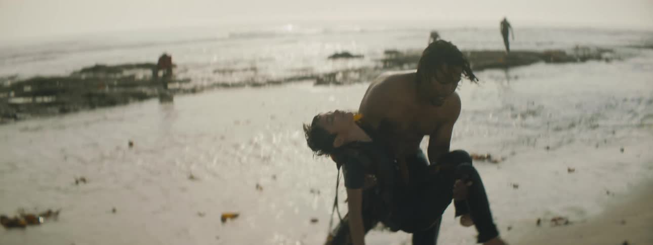 A man carries a woman to the beach from the ocean water and starts pushing on her chest in a cpr motion