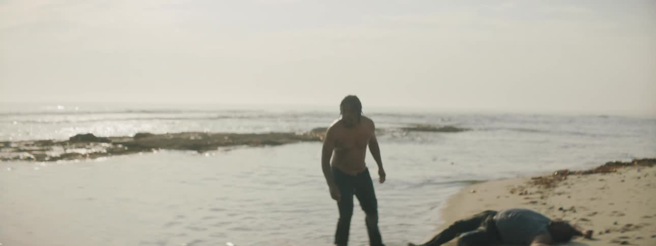 A man helps a person walk from the ocean water and they collapse on the beach next to other people laying in distress