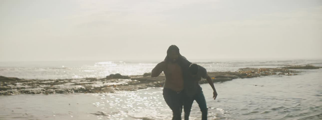 A man on the beach rescuing another man and putting him on the sand with the sky and clouds in the background