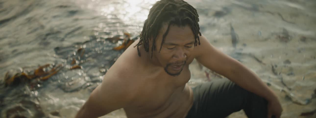 A shirtless man catches his breath while sitting in the shallow water of a beach before rising and walking away