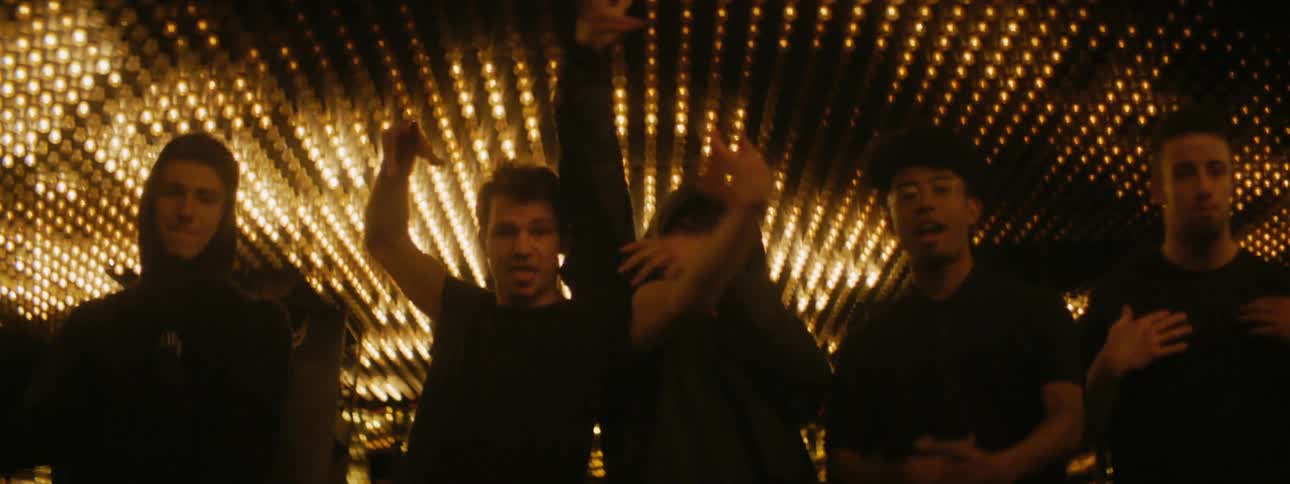 A group of teenage boys stand under bright lights and dance