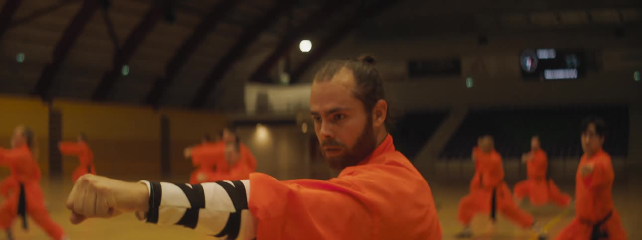 Men and women in orange outfits are performing martial arts moves in unison on a gym floor