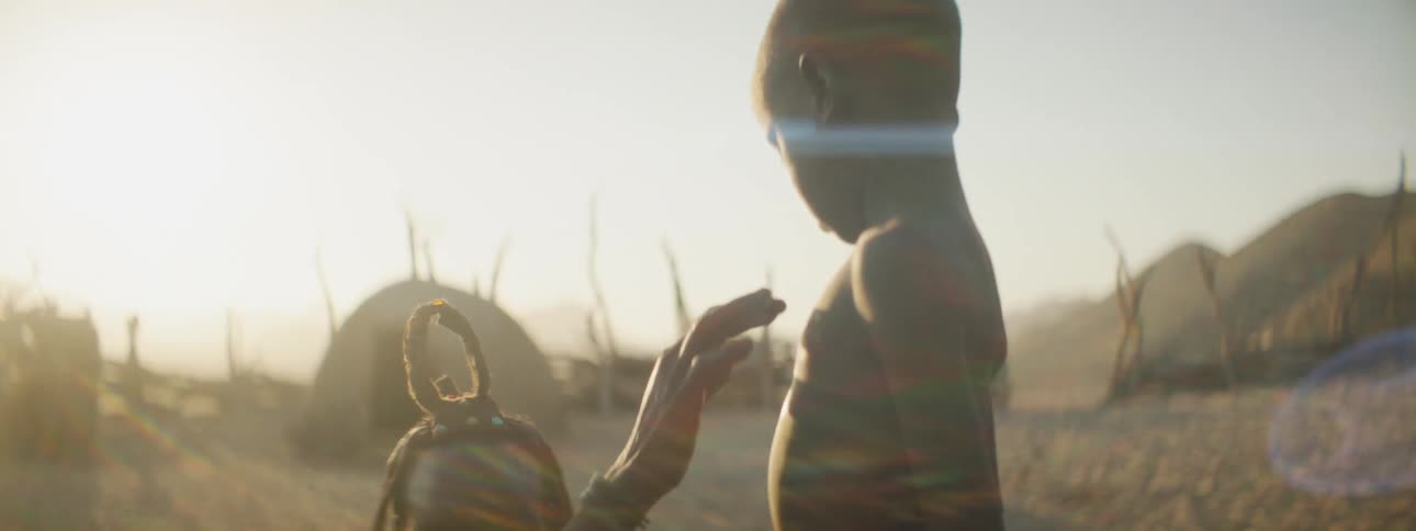 Mother places her hand on sons chest then has him turn around and places her hand on his back they are in the sand in front of huts that have large sticks around them in front of the sun and blue sky