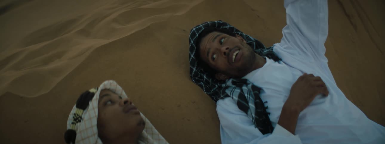 A man talks smiles and gestures upward while lying on sand dunes near another man