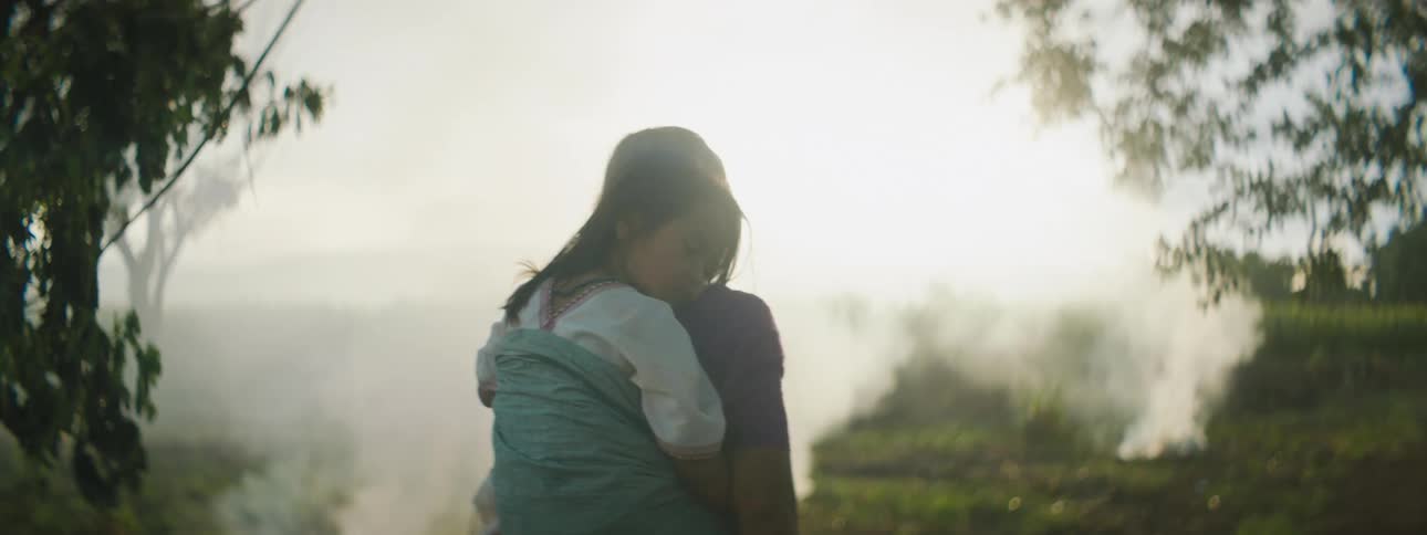 Grey haired grandmother carries granddaughter in sling past smoking fires set in green fields
