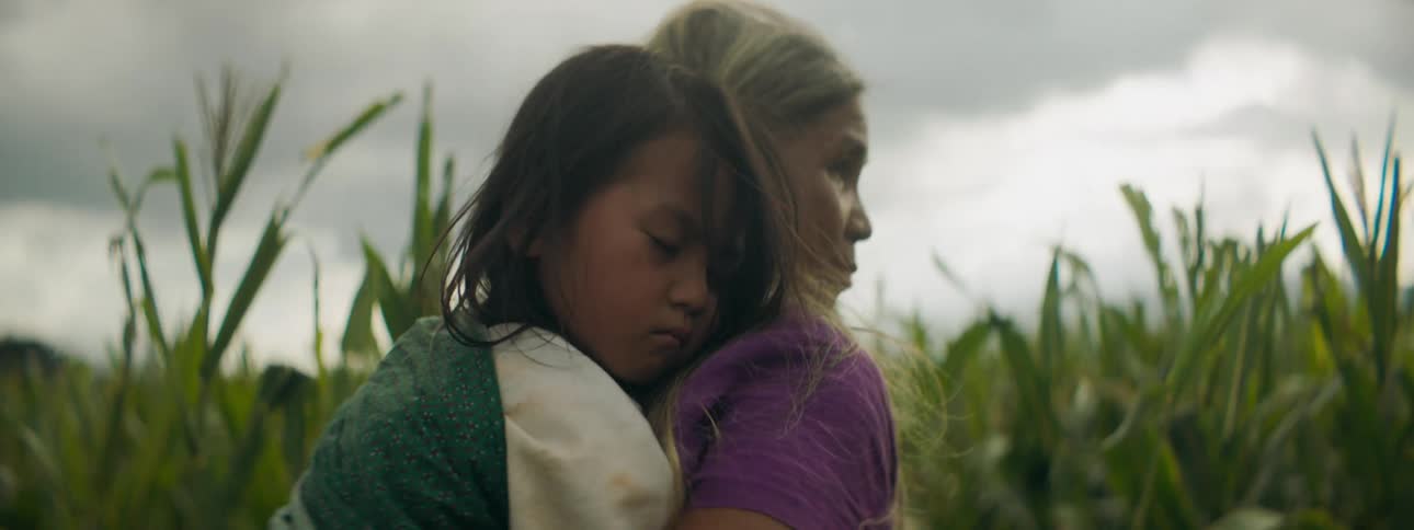 A woman walking in a cornfield with a girl on her back with the sky and clouds in the background