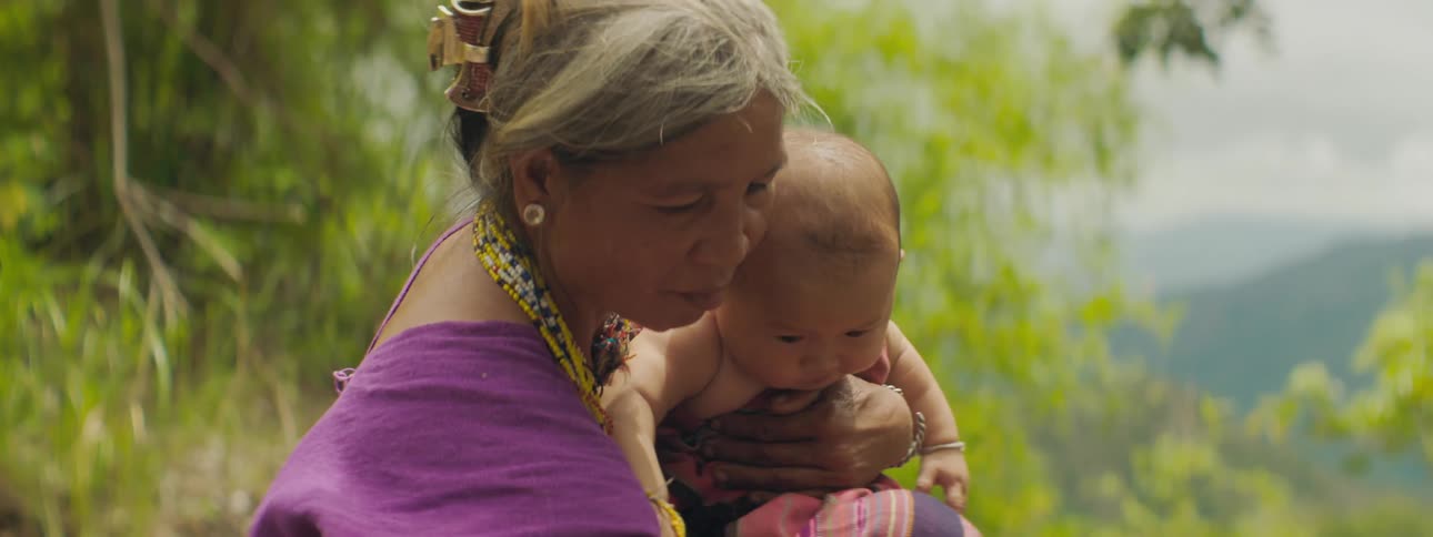 Grandmother holds baby near vegetation with mist covered mountains in background