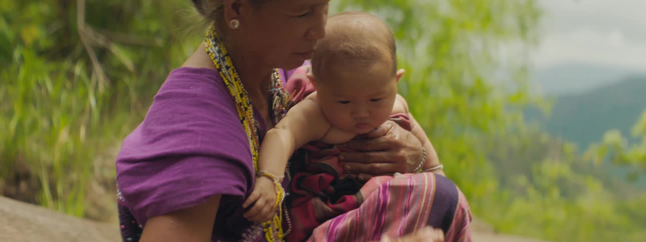 An elderly woman holds a baby in her arms and rubs water on its forehead