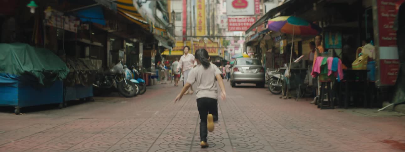 A little girl runs down a pathway with shops and pedestrians