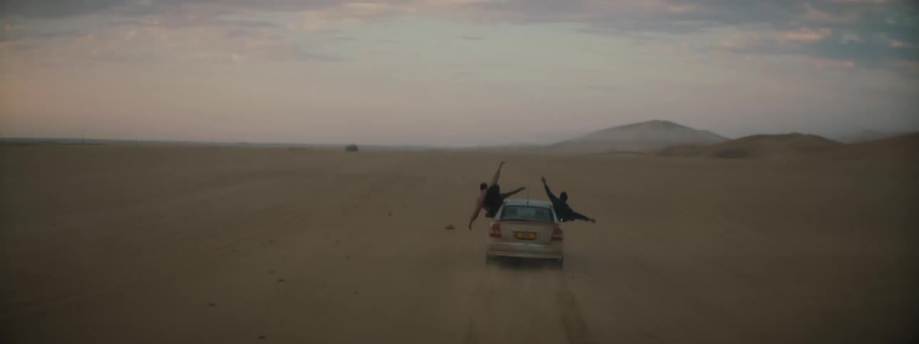 The driver and two passengers in a tan sedan hang out of the windows with their arms raised as it drives through a desert