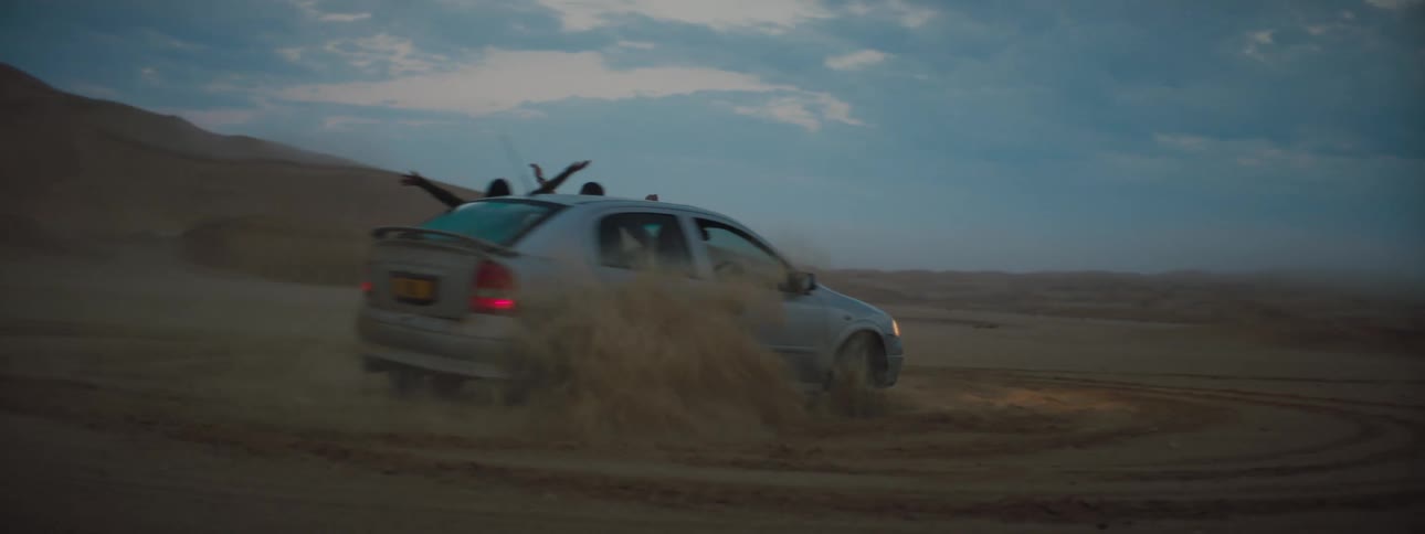 Two people hang out of car windows as it does donuts in a desert terrain