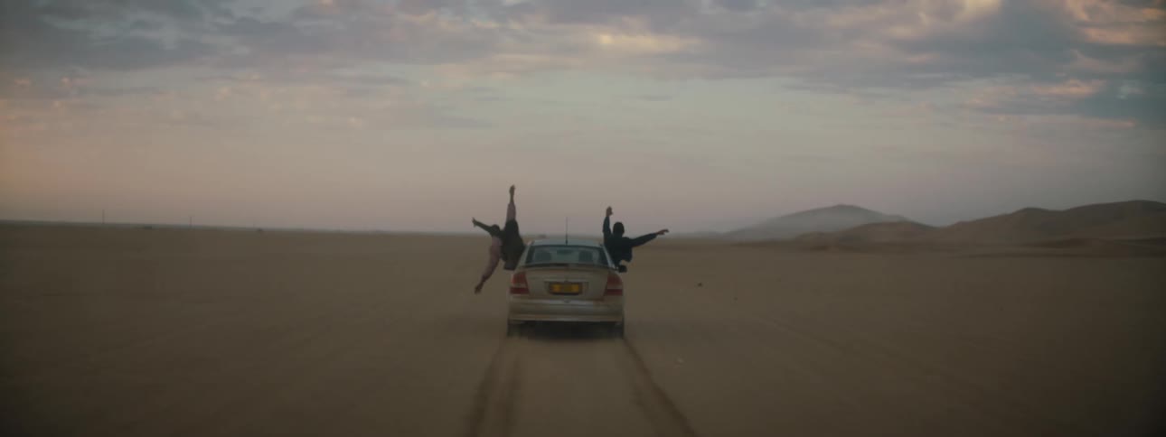 People hanging out of car windows as it drives across a desert