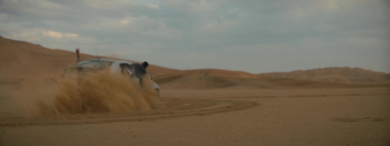 Two people hang out of a vehicle as it does donuts in a dry desert terrain
