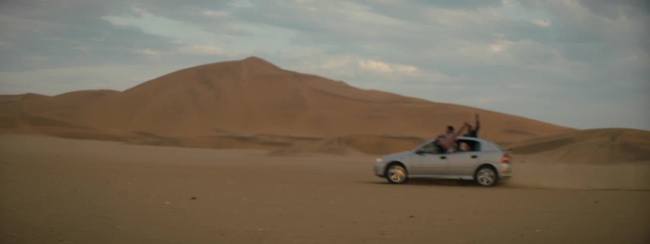 People standing through open car windows while driving across a sandy desert