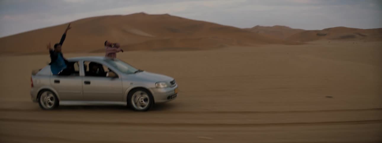 Two people hang out of the car windows as it drives over a dry desert terrain