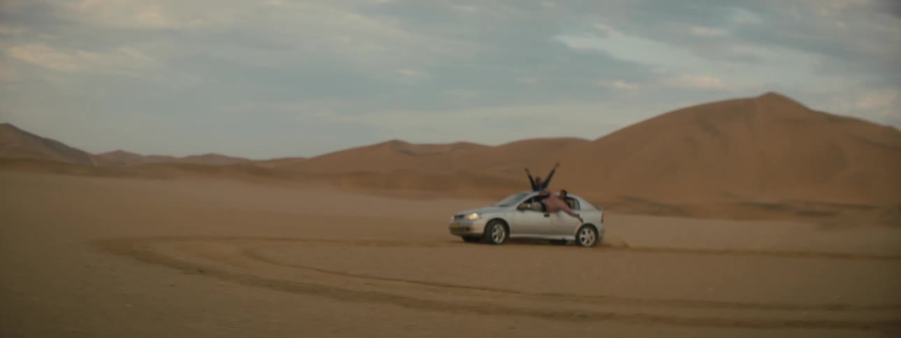 Two people hang out of a vehicle doing donuts in a dry desert terrain