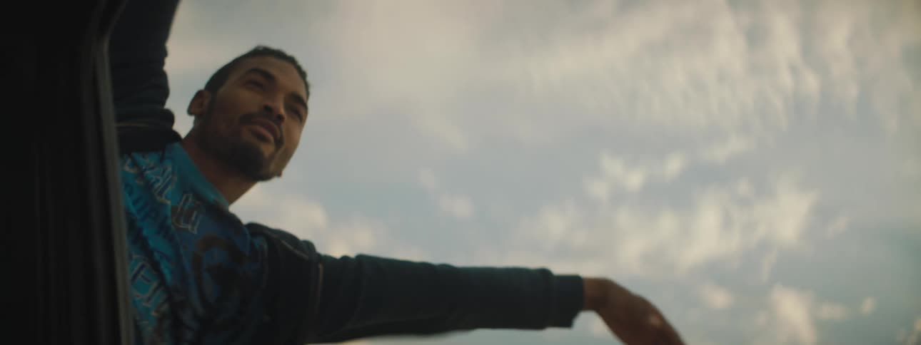 A man hangs halfway out the backseat window of a moving vehicle and waves his arm