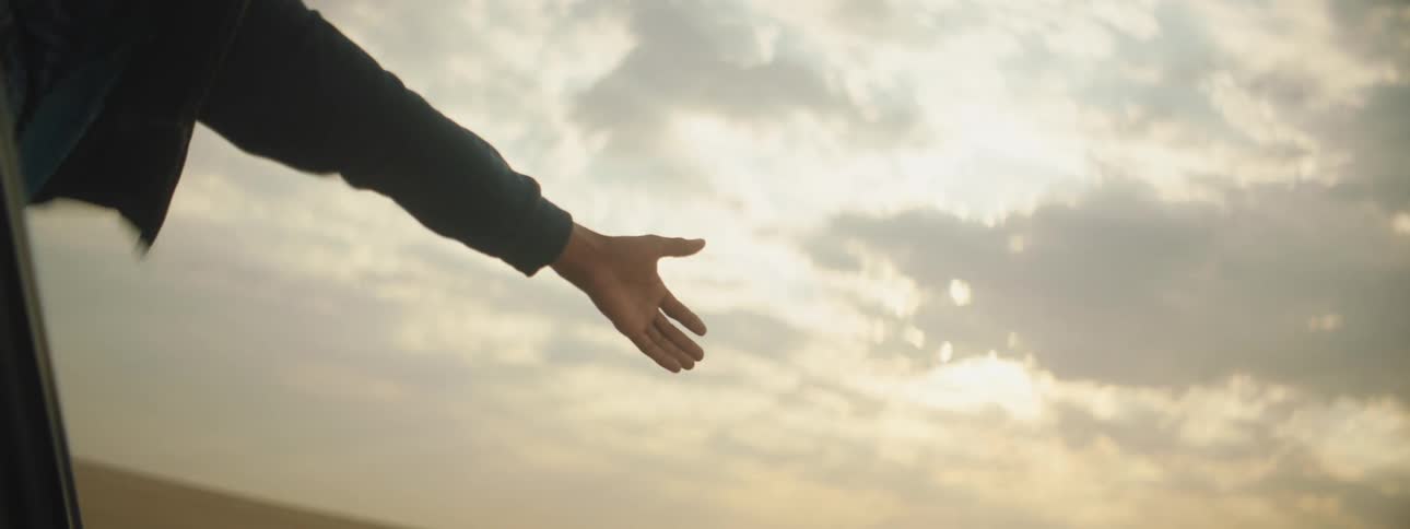 A person leans out of the window of a vehicle with their arms held wide as the vehicle travels through the sands of a desert