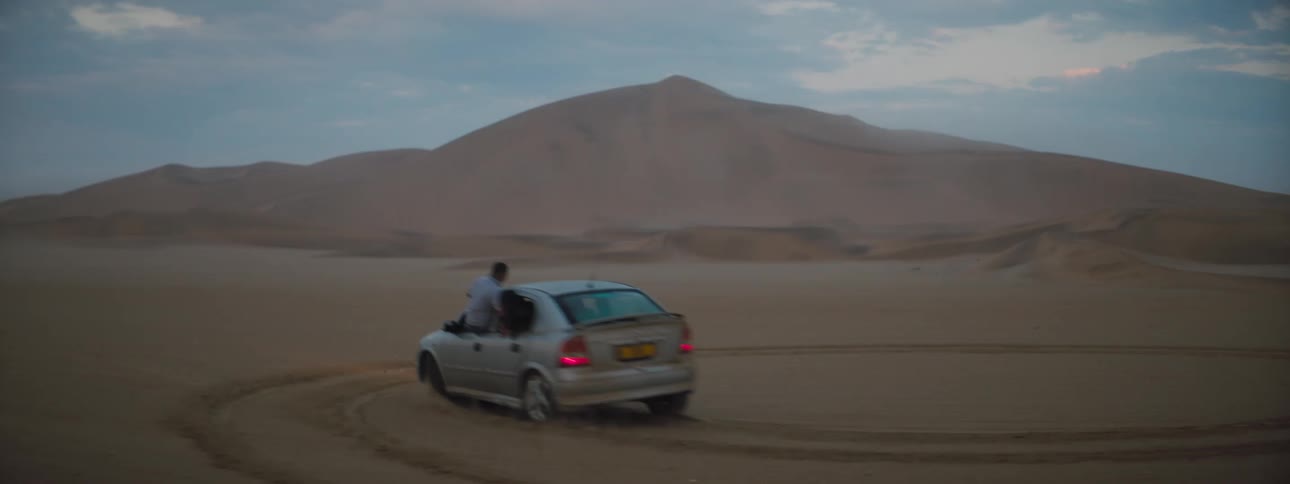 A car spins around doing donuts in a desert terrain with two people hanging out of the car windows