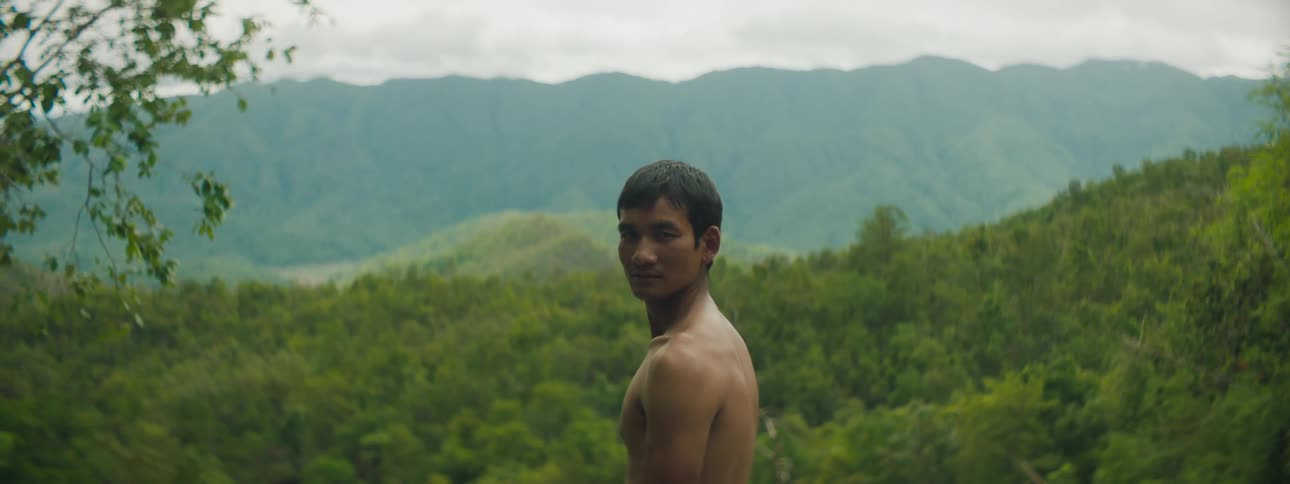 A shirtless man looking out over a forested countryside turns to look into the camera behind him