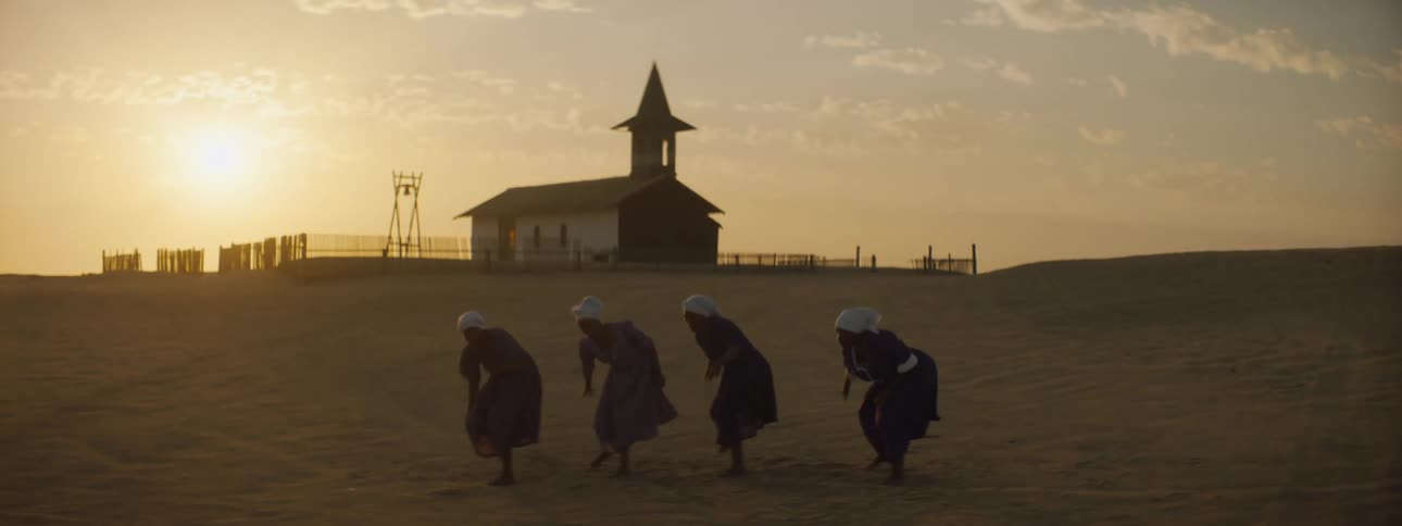 Four women wearing dresses dance in unison in front of a church at dusk outside