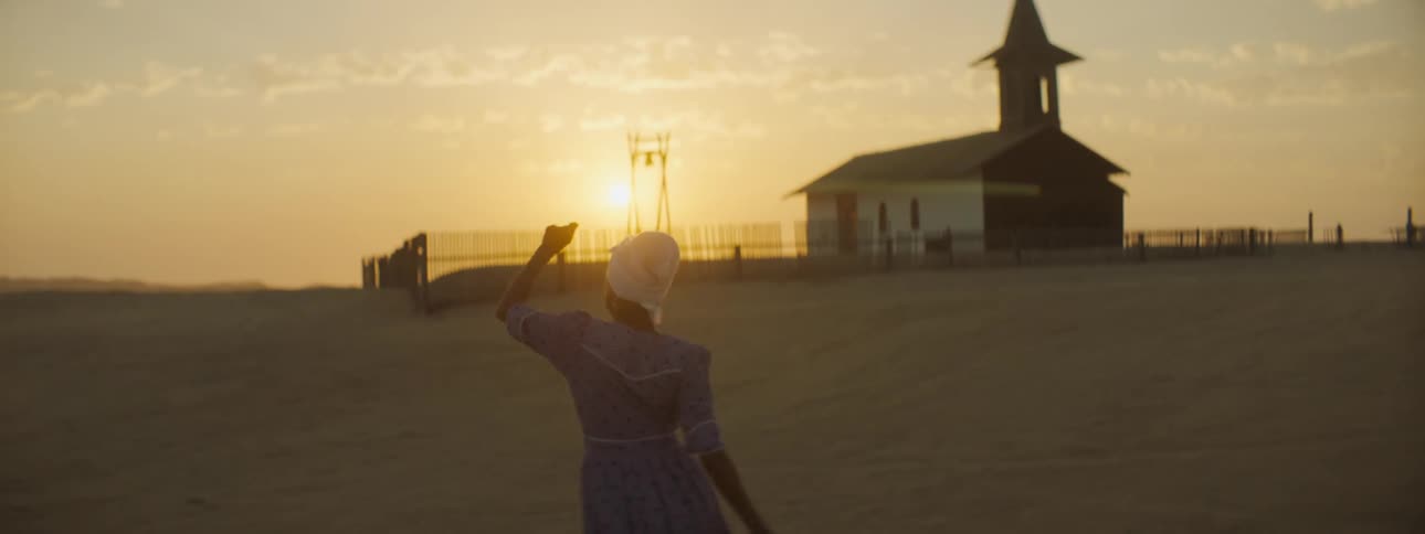 Sun sits above tinted sky as woman dances across sand to building sitting in fenced area