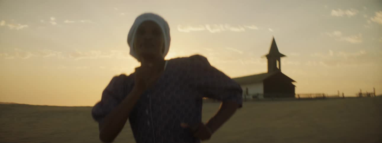 Woman in dress and white head covering dances in the sand at sunset with a church and fence in the background with a blue and orange sky with white fluffy clouds