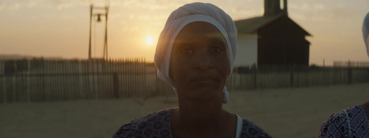 Women stand outside of a remote village with a fence and house in background while the sun sets behind them