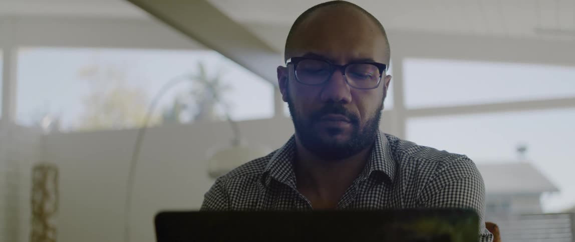 A man sits at home and rubs his chin as he looks at his laptop