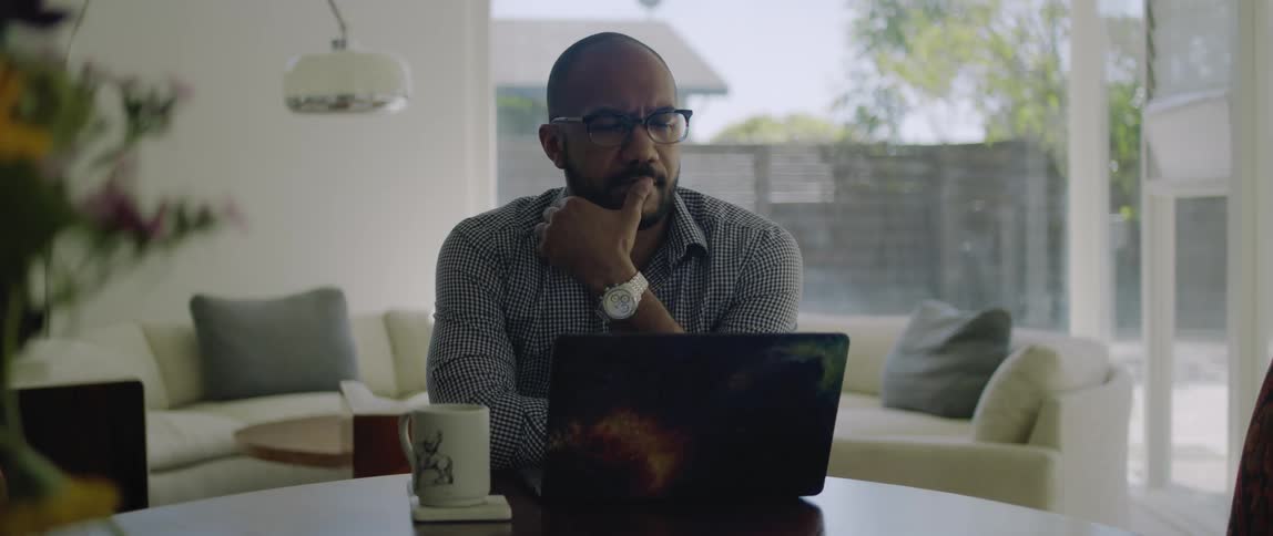 A man using a laptop at a table in the living room of a house stops to think for a moment