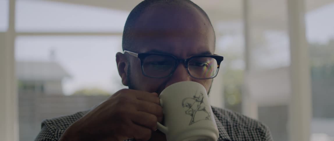 A man wearing glasses slowly takes a sip from his coffee cup as he sits indoors