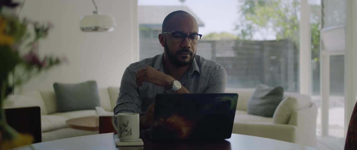 A man is sitting in his home with a cup of coffee and looking at his laptop