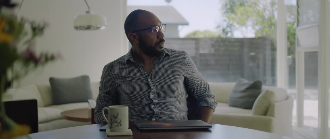 A man sitting at a table puts his glasses on then grabs his phone as he stands to walk away