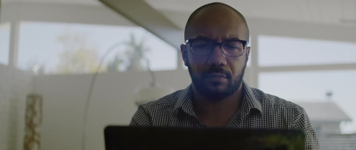 A man wearing glasses looks down at his laptops screen as he types