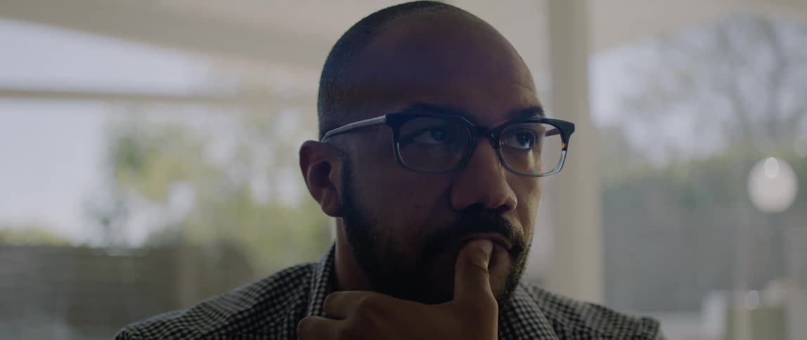 A man presses his mouth with his thumb while lost in thought as he sits indoors during the day
