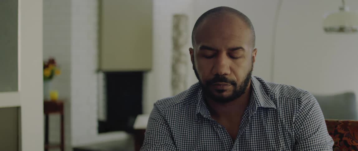 A man sitting at home looks to the side then goes back to looking down at something in front of him