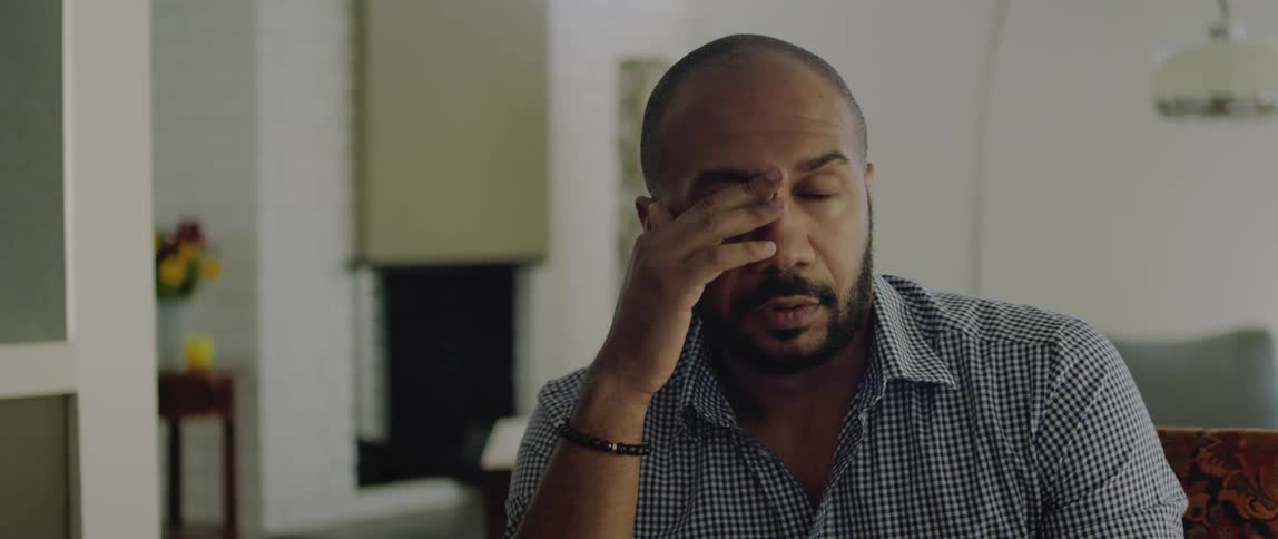 A tired man sitting in a room rubs his eyes and looks to the side