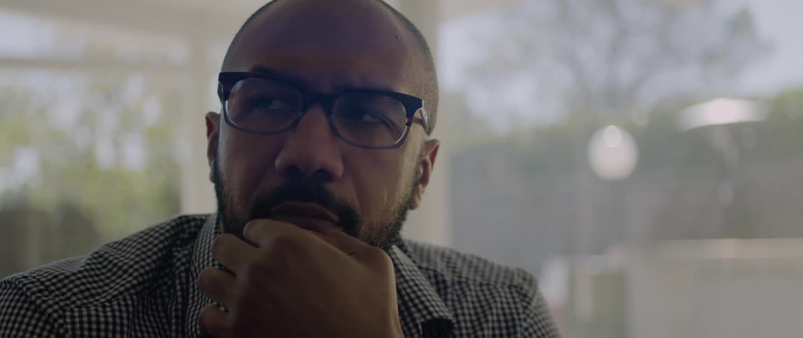 A man wearing glasses is looking around with his hand on his face