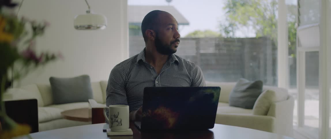A man is sitting at a table with a laptop computer and a coffee cup and he puts on his glasses and starts typing on the keyboard