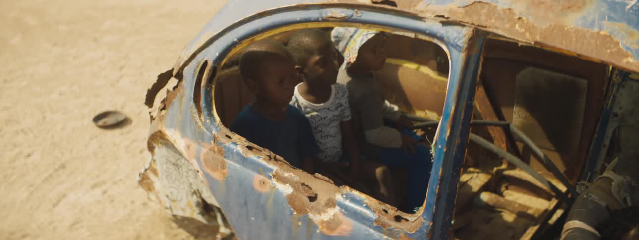 Children play in wrecked blue car sitting on sandy ground