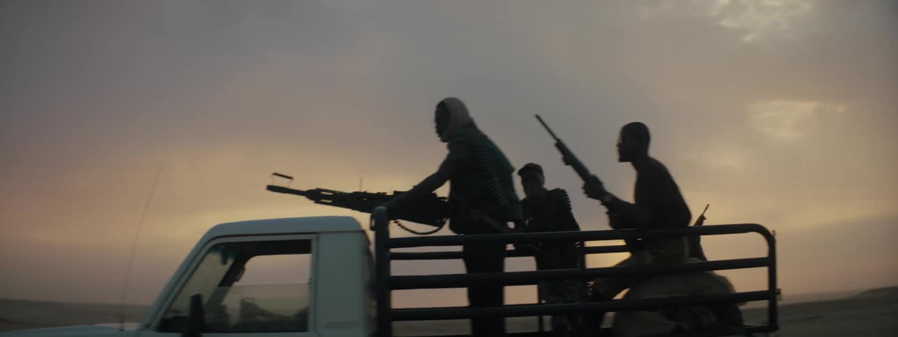Four men riding in the back of a truck with a machine gun and rifles leaning against railing on the bed of the truck behind them is an open field or grass plain and the orange purple blue and pink of the sunset