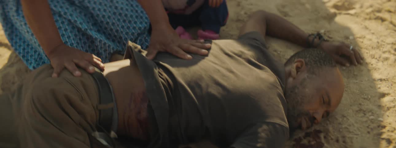 Man lays dead in the sand while a woman has her hands on his back the man who shot him is standing above them looking down at the dead man there are buildings and a blue truck in the background