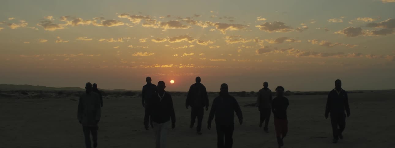 A group of black men walk together through a desert with the setting sun at their back